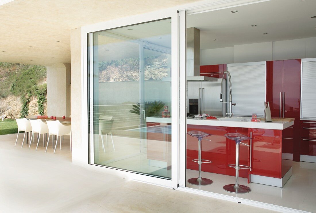 Designer-Barhocker an Küchenblock mit roter Front, gesehen durch offene Schiebefenstertür von der Terrasse eines modernen Ferienhauses