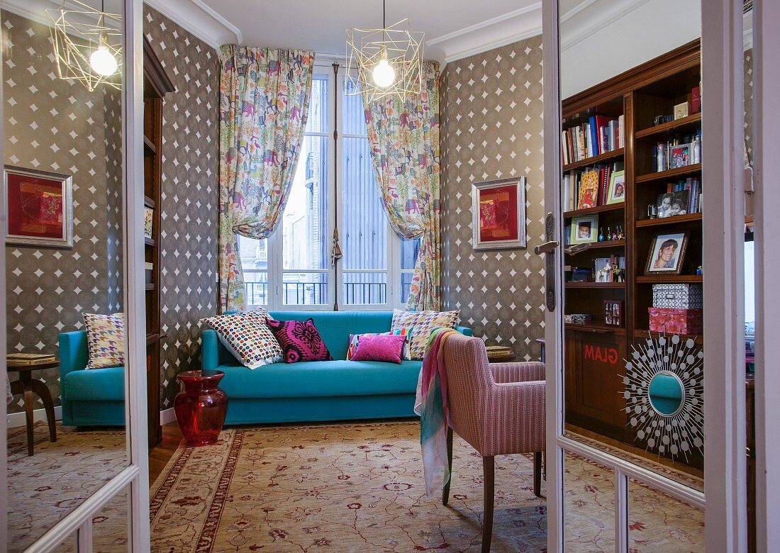 Light blue couch in front of window with draped curtains and patterned wallpaper in eclectic interior seen through open double doors