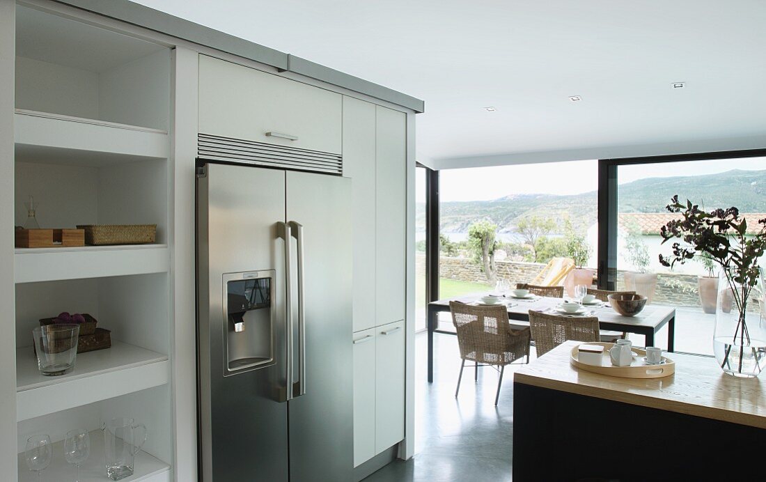Doppeltüriger Kühlschrank aus Edelstahl, flankiert von Küchenschränken und Regal; im Hintergrund Essplatz vor Panoramafenster