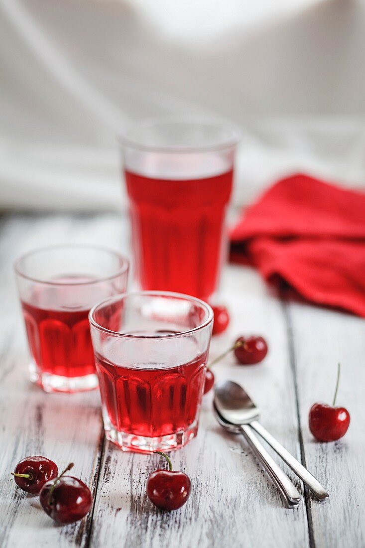 Three glasses of cherry jelly, fresh cherries and tea spoons on a wooden surface