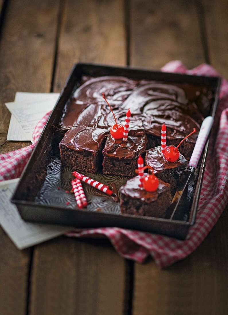 A chocolate-cinnamon tray bake cake with a buttermilk glaze cut into slices and decorated with candles and glacé cherries