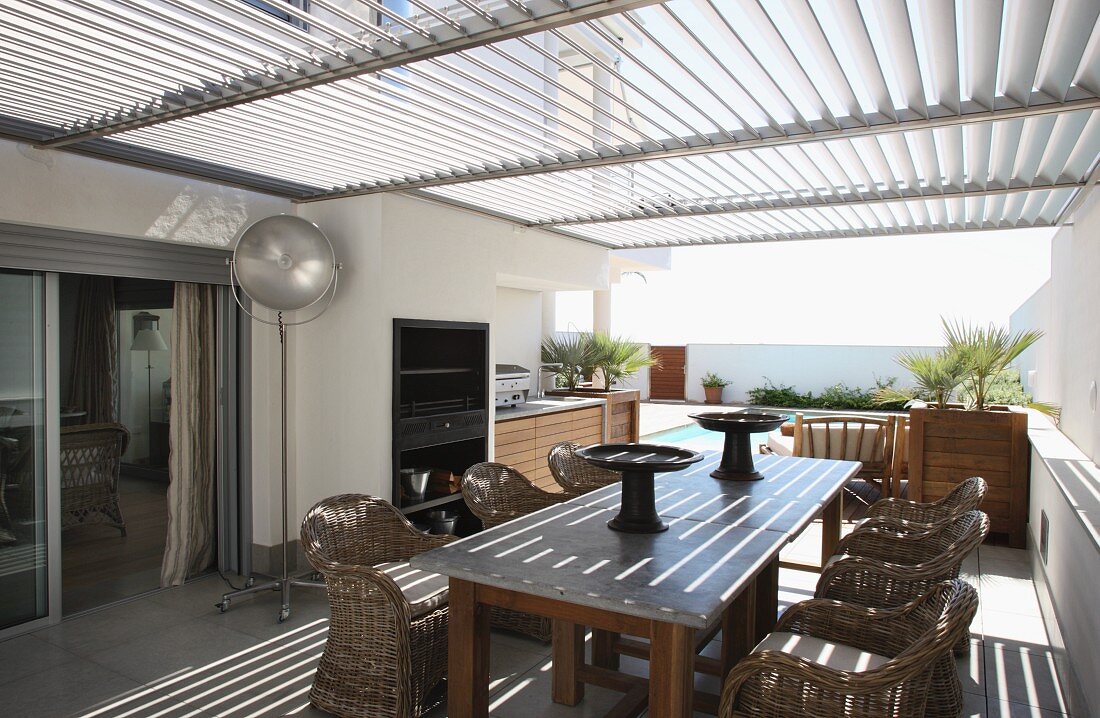 Rattanstühle um Holztisch auf mit Sonnenschutzlamellen überdachter Terrasse