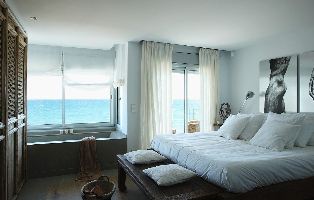 Double bed with white bed linen and bathtub below window with panoramic sea view