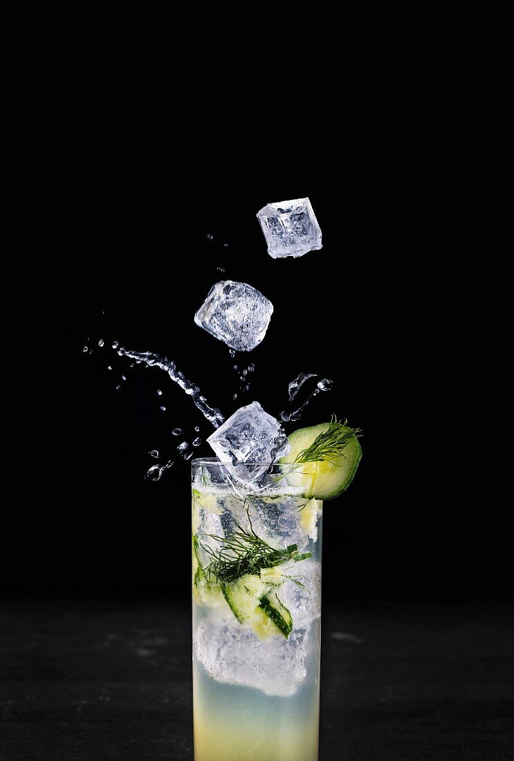 Ice cubes falling into a summer cocktail garnished with cucumber