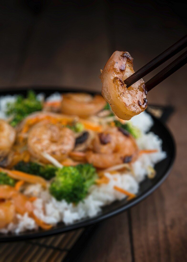 Prawn stir fry with rice, broccoli and carrots; in the foreground a glazed prawn between two chopsticks