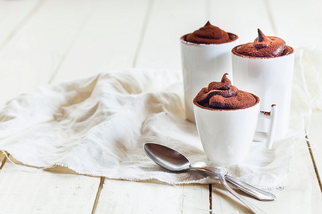 Three cups of vegan mousse au chocolat, coffee spoons and a cloth on a white wooden surface
