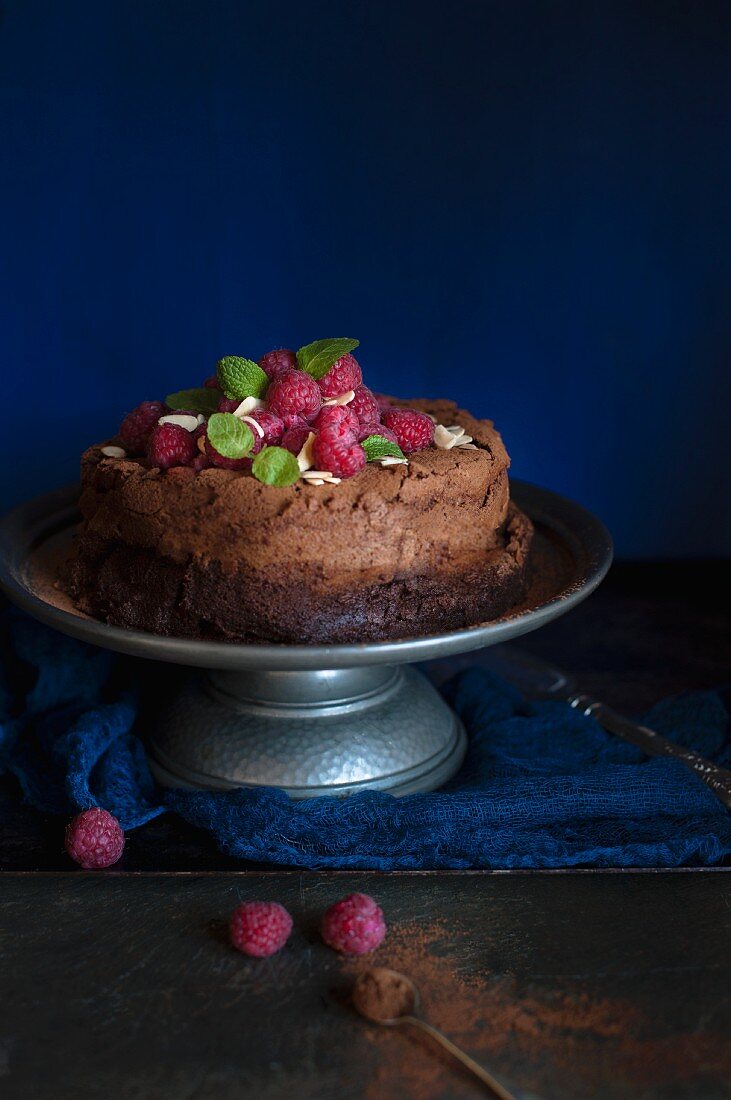 Dark chocolate cake on a metal cake stand with fresh raspberries and mint