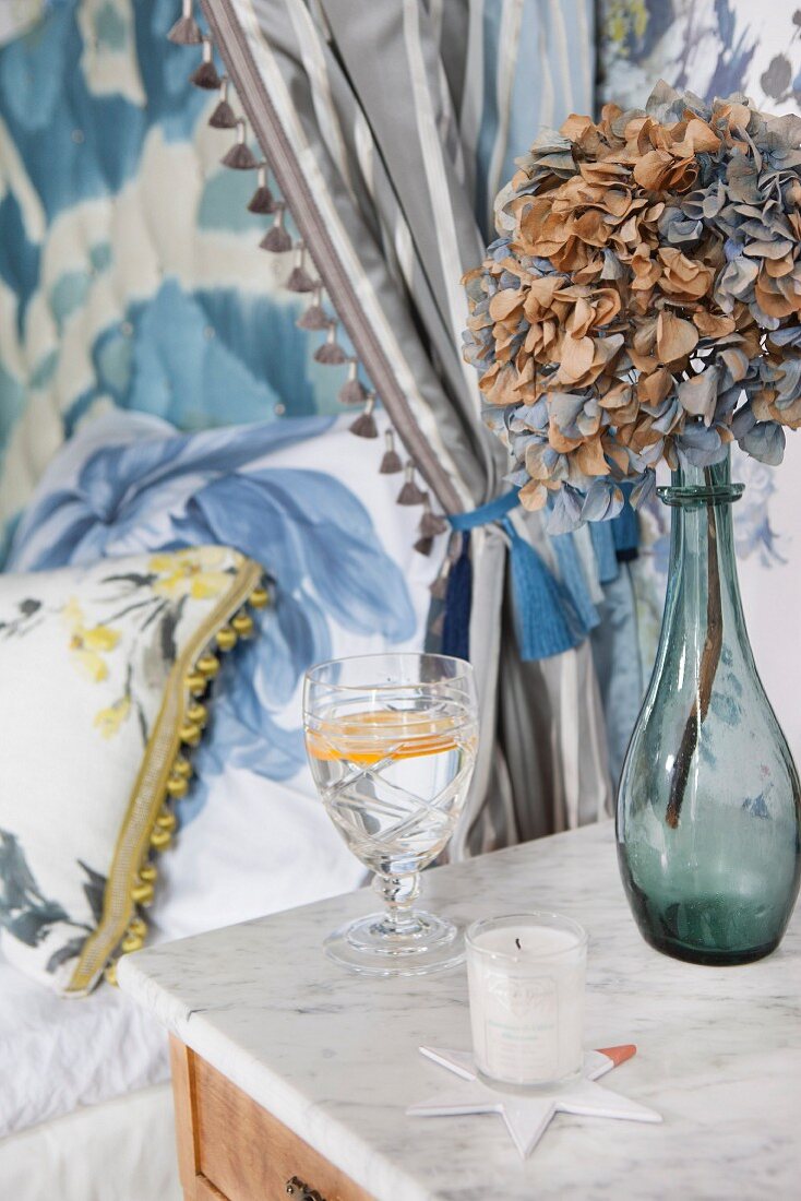 Getrocknete Hortensie in Glasvase und Kristallglas auf Nachttisch mit Marmorplatte, Bett mit blumigen Bezügen
