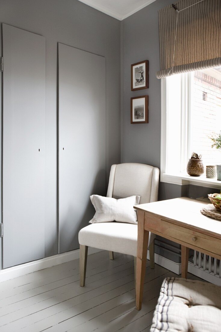 Kitchen table and upholstered chair below window next to grey-painted cupboard door