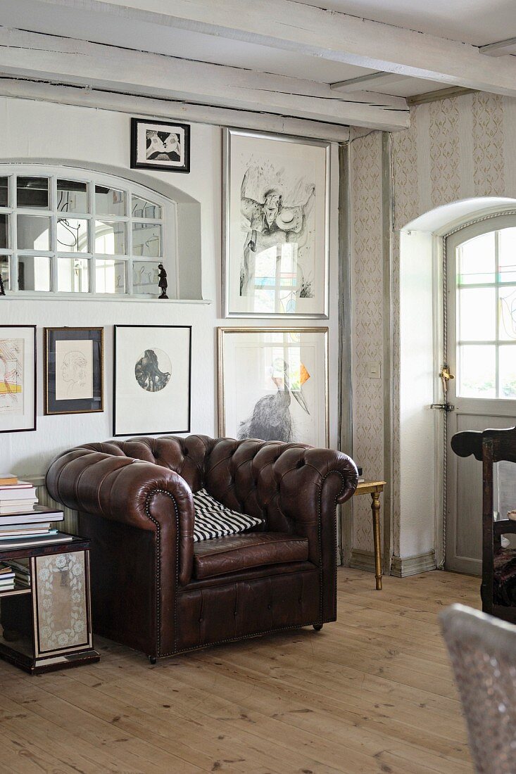 Brown leather armchair below collection of artworks on wall in rustic interior