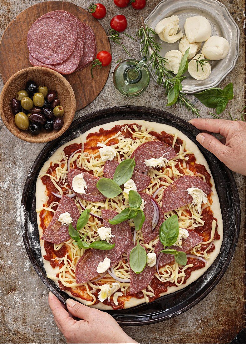 Hands holding an unbaked pizza with sausage and cheese surrounded by ingredients