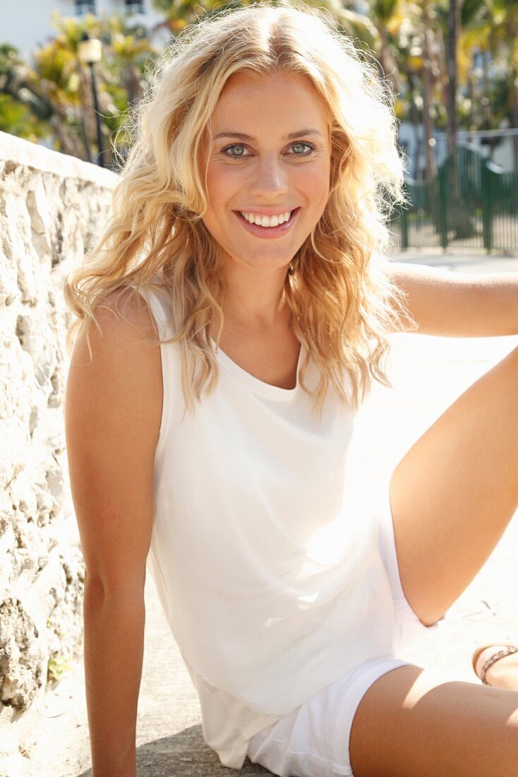 A blonde woman sitting against a stone wall wearing a white top and shorts