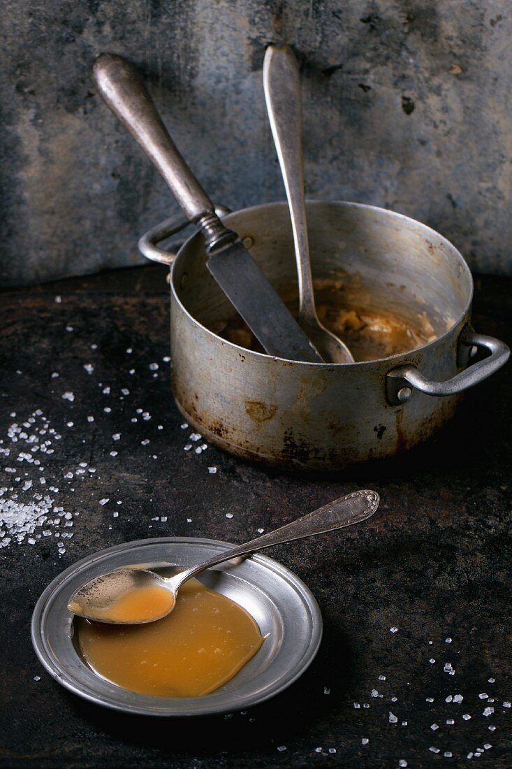 Selbstgemachte Karamellsauce auf Teller mit Löffel, verstreuter Zucker und alter Kochtopf
