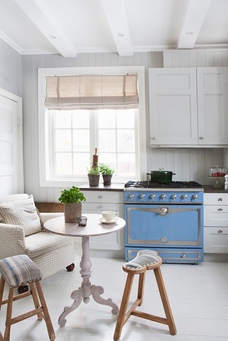 White, Scandinavian country-house kitchen with pale blue gas cooker, armchair, table and wooden stools in vintage ambiance