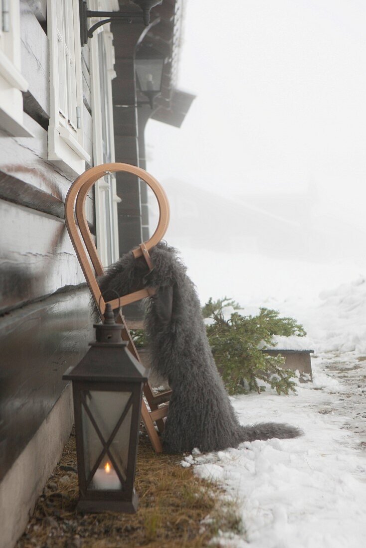 Bodenlaterne mit brennender Kerze neben Schlitten mit Tierfell an Holzhaus