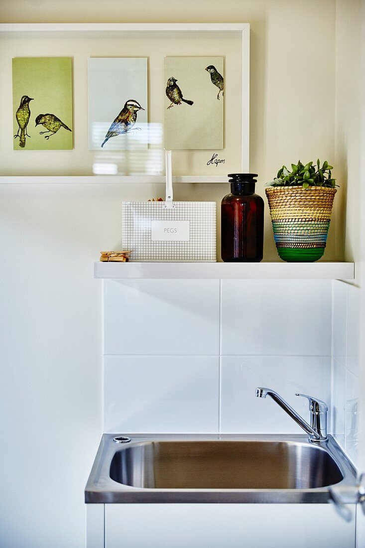 Stainless steel sink against white-tiled wall below shelf and framed triptych of birds on painted wall
