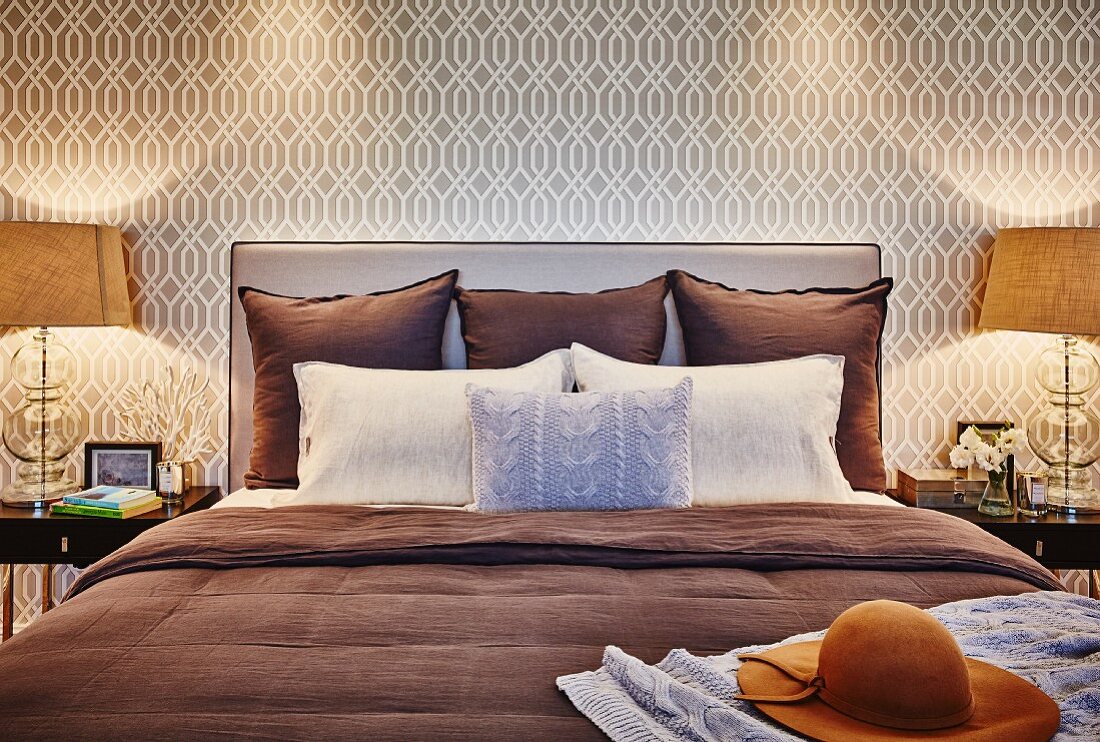 Pillows and cushions on double bed with upholstered headboard against wallpaper with beige retro pattern