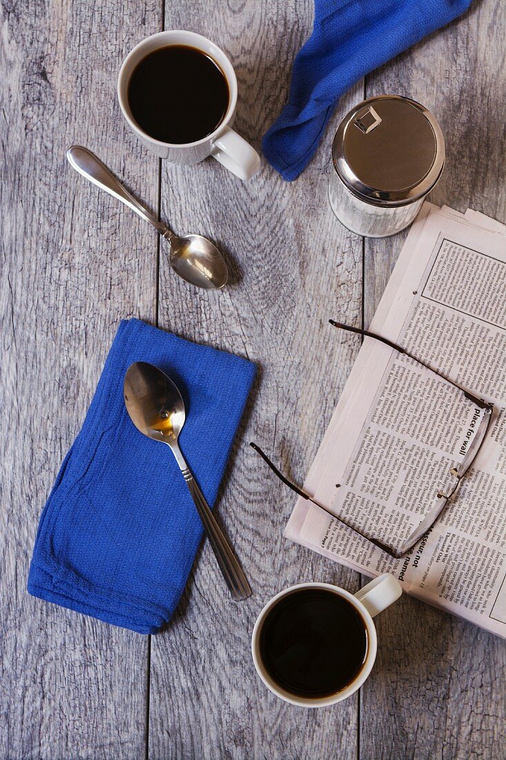 Frühstückstisch für Zwei mit Kaffeetassen, Löffel, Servietten, Zuckerdose, Morgenzeitung und Brille