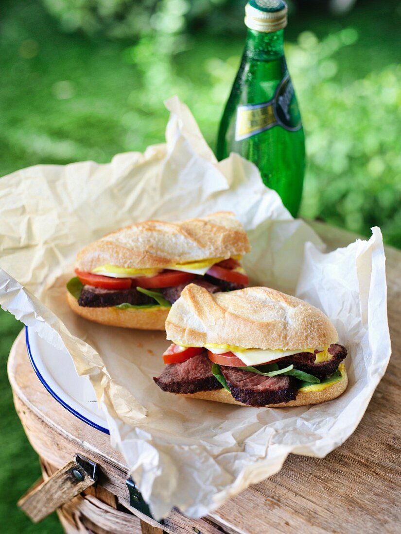 A grilled steak sandwich with mustard, cheese, tomatoes and spinach in parchment paper on a picnic basket