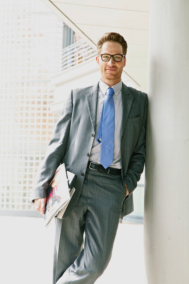 Mann in grauem Anzug und Unterlagen unter dem Arm lehnt an Säule vor Bürogebäude