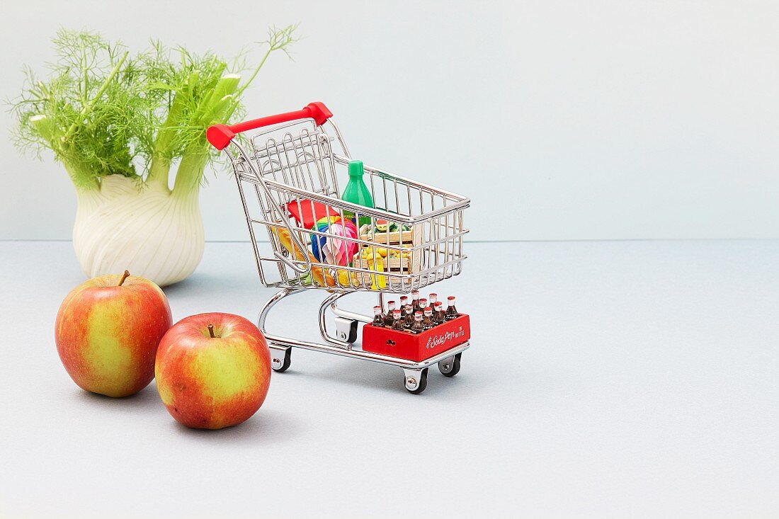 A mini shopping trolley filled with toy foodstuffs next to apples and a bulb of fennel