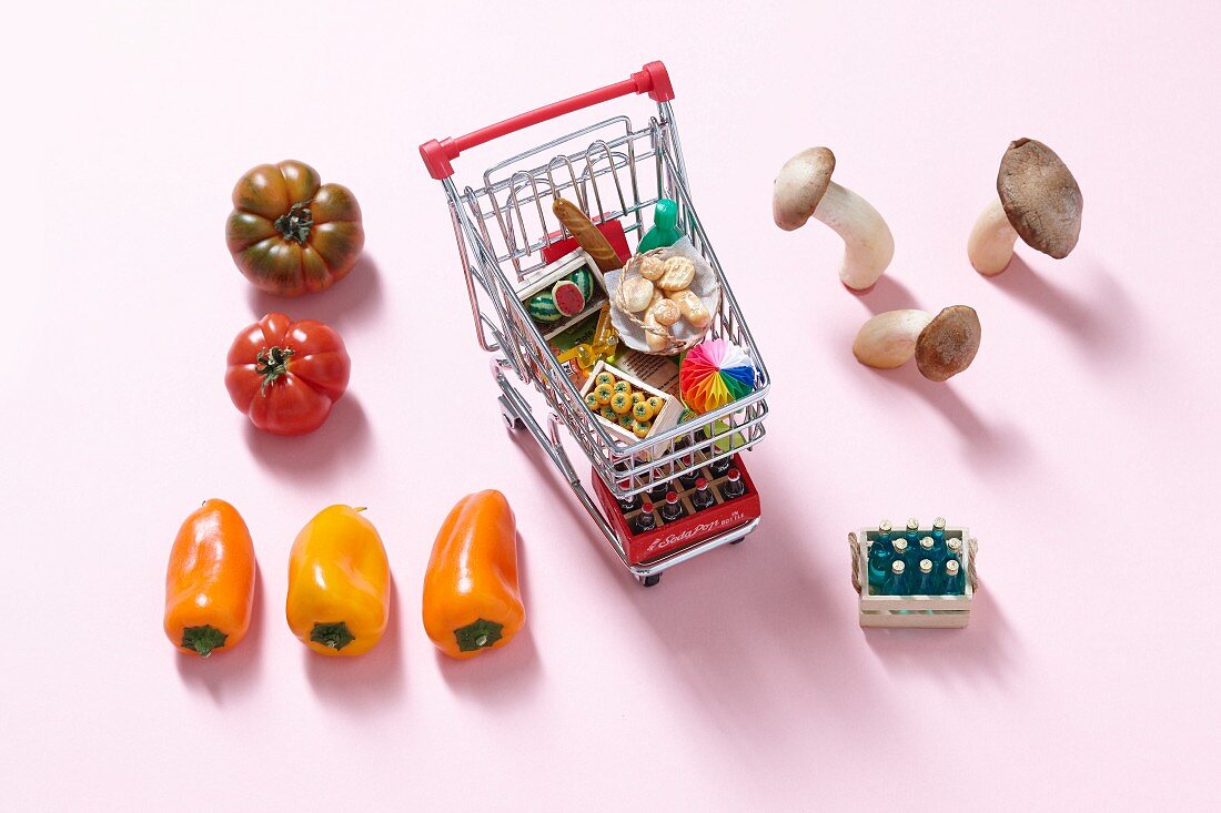 A mini shopping trolley filled with toy foodstuffs next to fresh vegetables and mushrooms