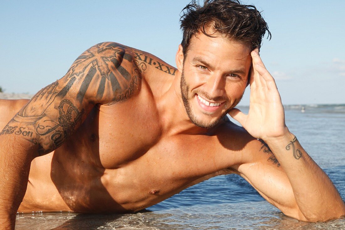 A young, topless, tattooed man lying on a sandy beach