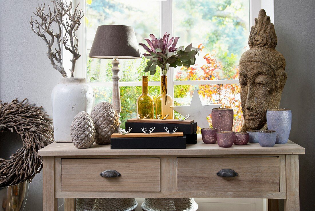 Verschiedene Vasen mit Blumen und Windlichter, seitlich Steinkopf in asiatischem Stil auf Holztisch vor Fenster