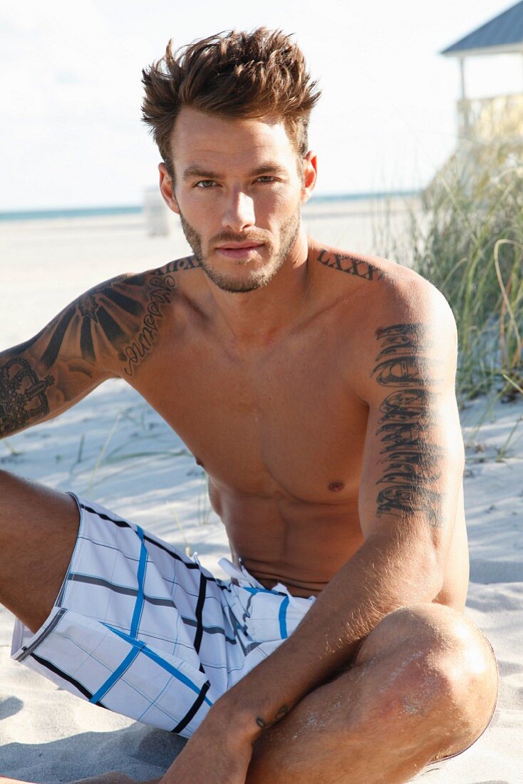 A young, tattooed man sitting in the sand wearing bathing shorts