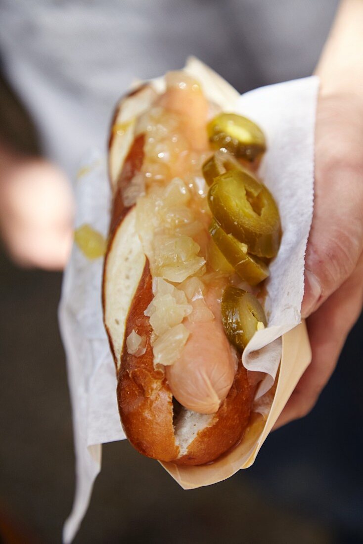 A hand holding a lye bread roll with a sausage, onions and gherkins