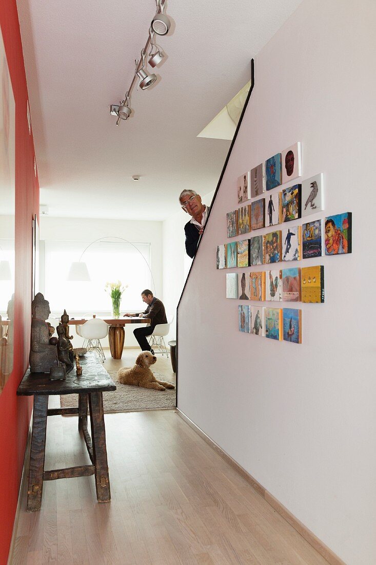 Rustic wooden table opposite gallery of pictures on wall of staircase; man walking up stairs and man sitting at dining table next to window