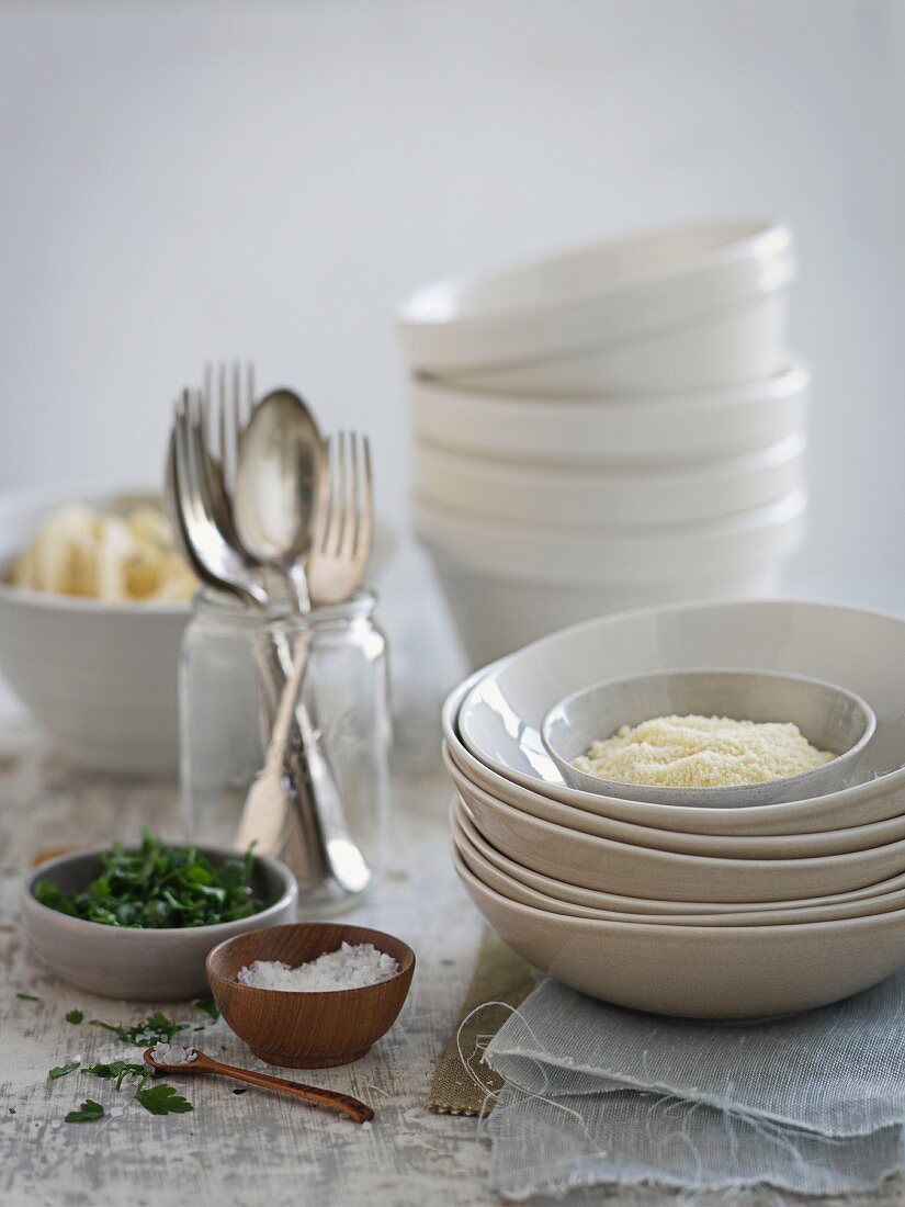 A stack of bowls, herbs, salt, grated cheese and cutlery