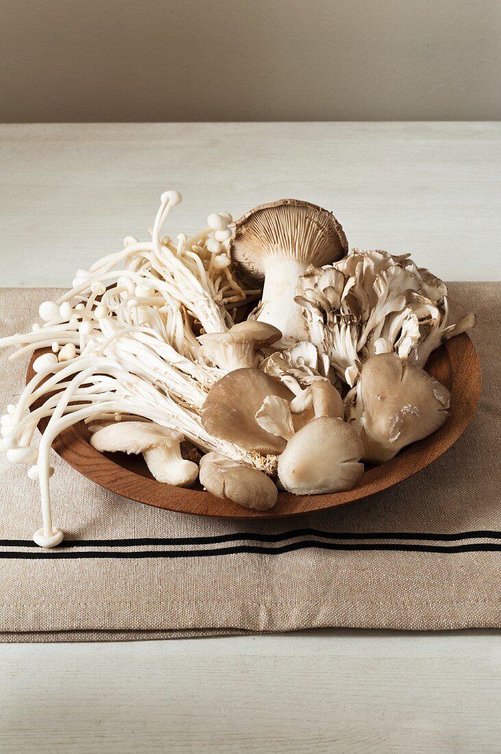 Various fresh mushrooms in a wooden bowl on a tea towel