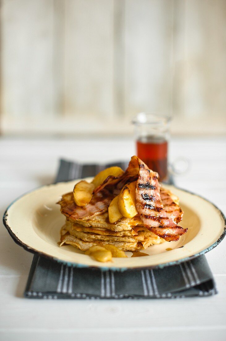 A stack of pancakes on a plate with grilled bacon and apple with maple syrup in the background