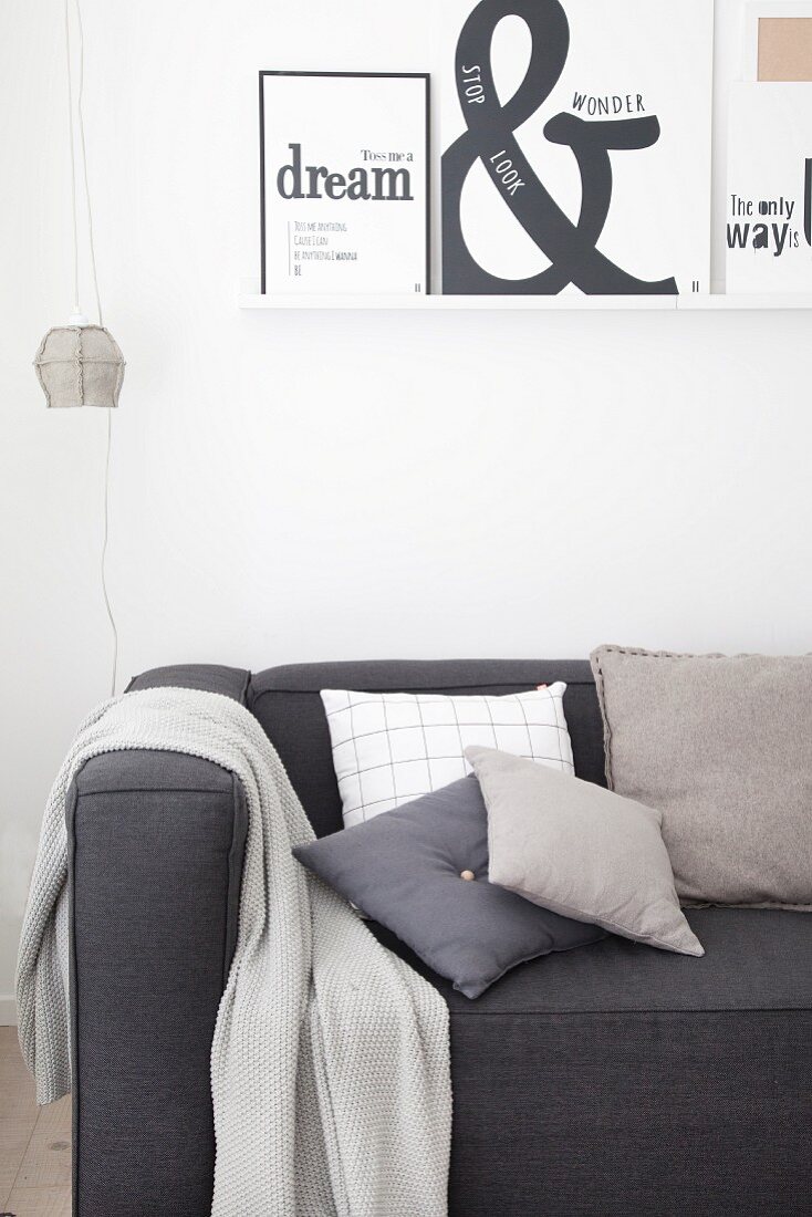 Various cushions and blanket on charcoal-grey couch below printed graphic artworks on white wall-mounted shelf