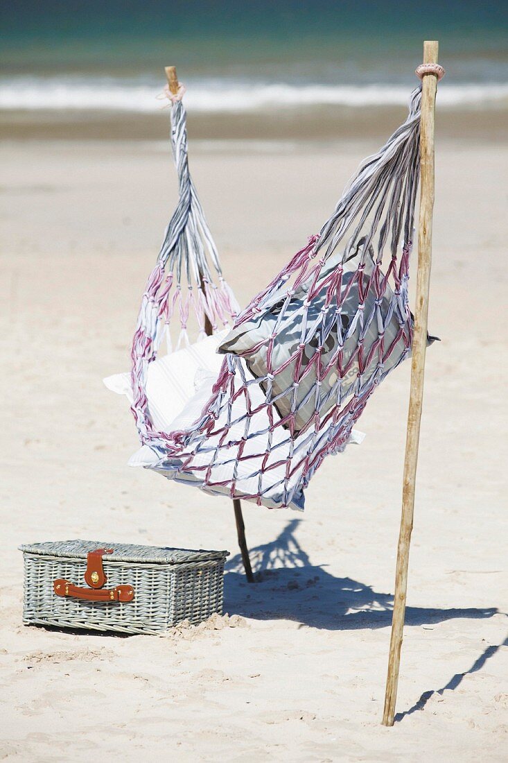 Hängematte auf Stöcken befestigt und in Sand gesteckt neben Picknickkoffer