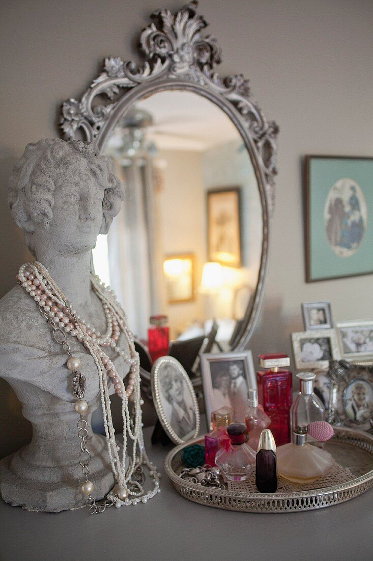 Familienfotos, Parfumflakons und Büste als Schmuckhalter auf Kommode vor dekorativem Silberspiegel