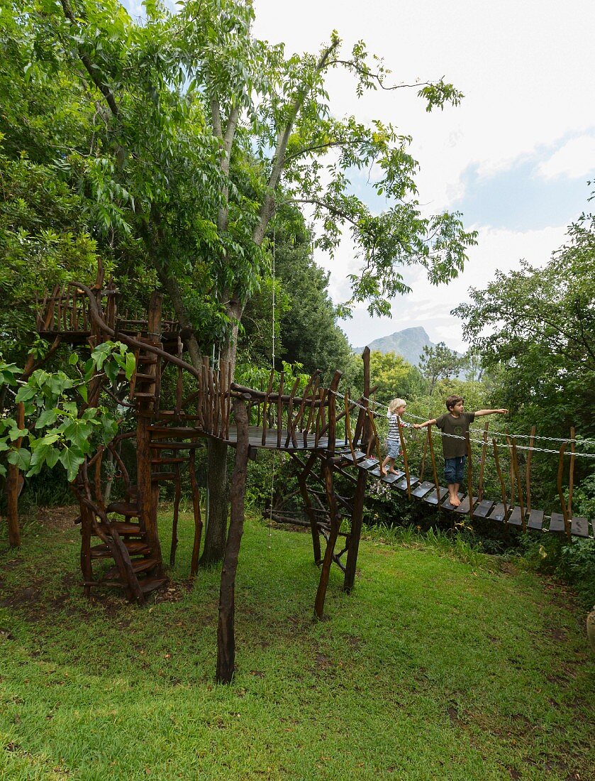 Two children playing in tree house with suspended bridge, tree-trunk platforms and spiral stairs in garden