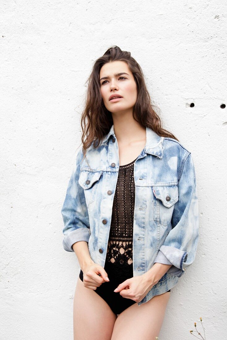 A young woman standing against a white wall wearing a bodysuit and a denim jacket