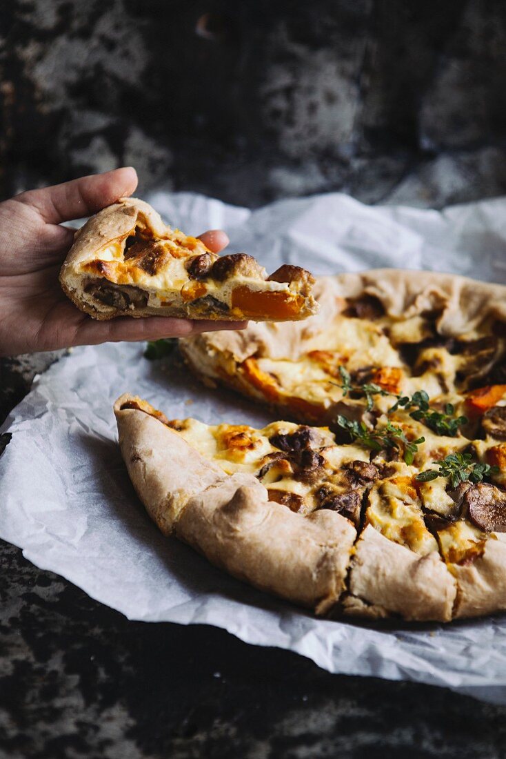 A hand holding a slice of ricotta tart with pumpkin and porcini mushrooms