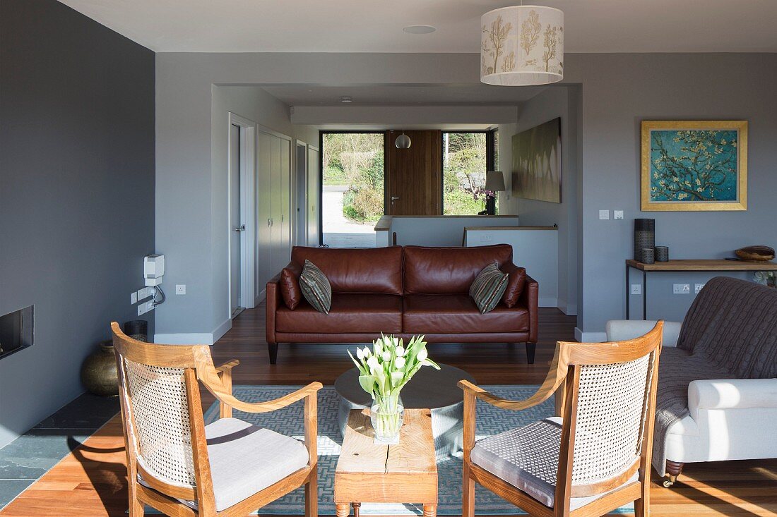 Cane chairs with seat cushions in sunlight and brown leather couch in modern open-plan interior with view into foyer