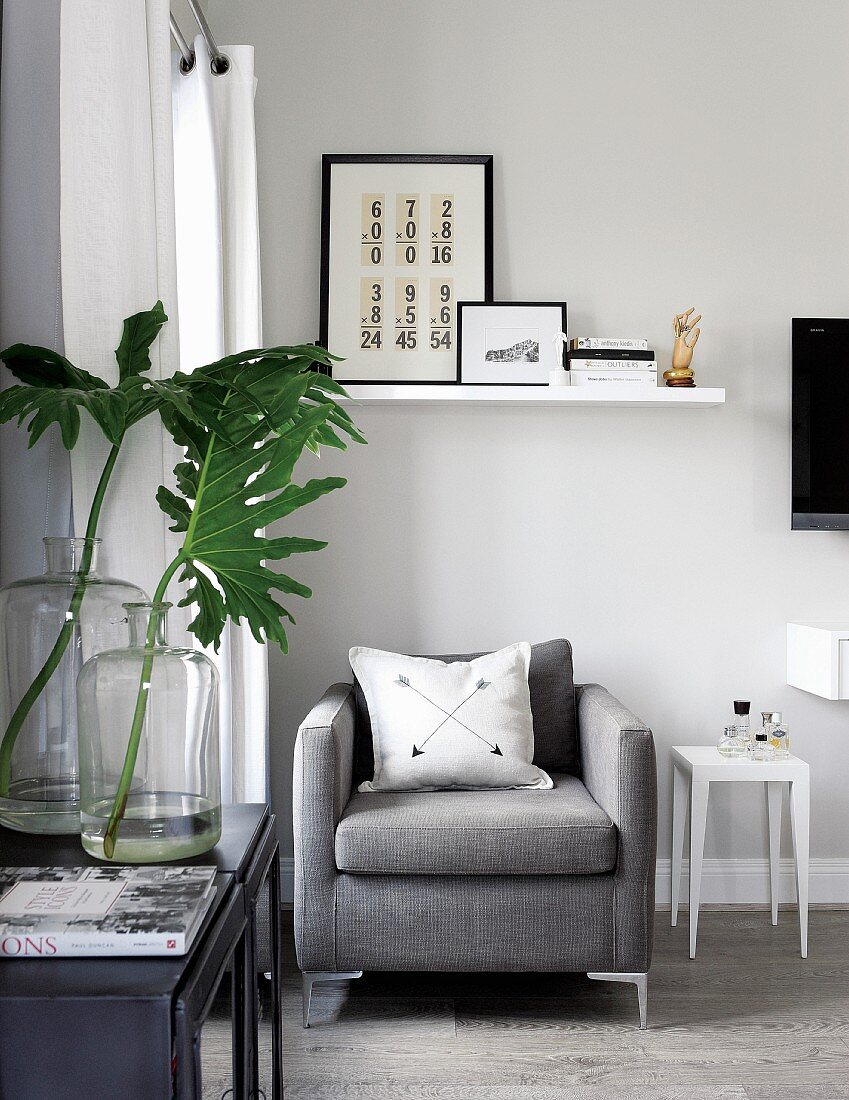 Grey armchair with scatter cushion under floating shelf and green leaves in vintage glass bottles in elegant interior