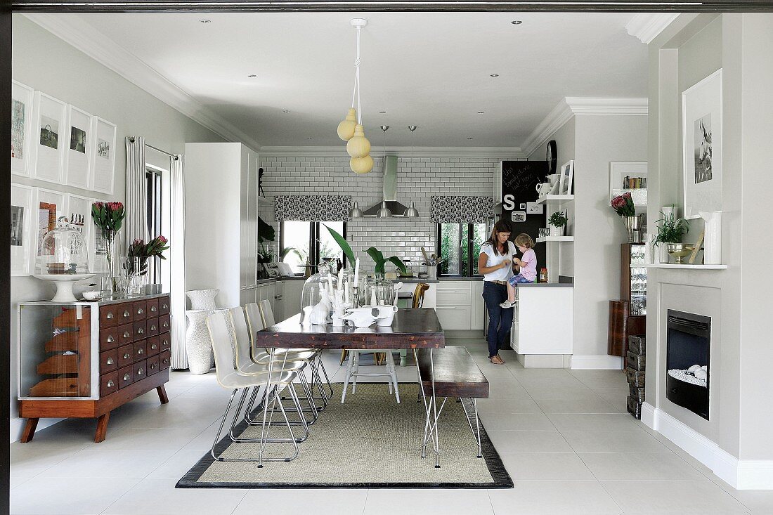 Dining table with matching bench and white chairs in open-plan interior; family in kitchen in background