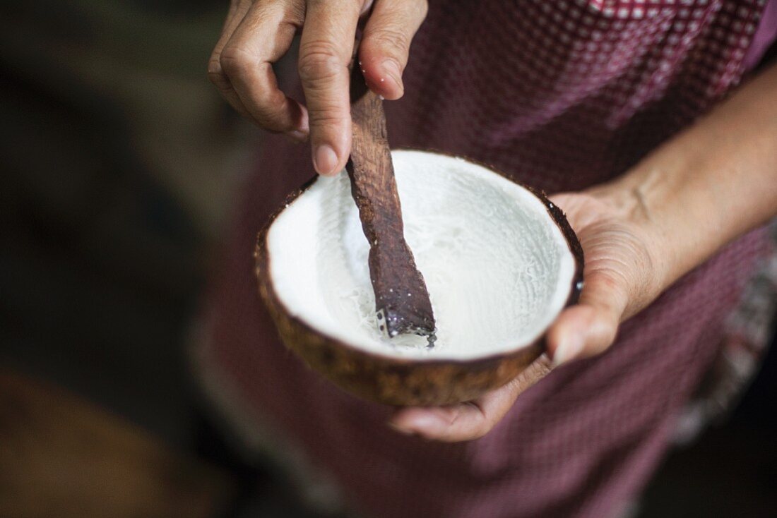 Coconut marrow being scratched out of a coconut shell with a wooden stick