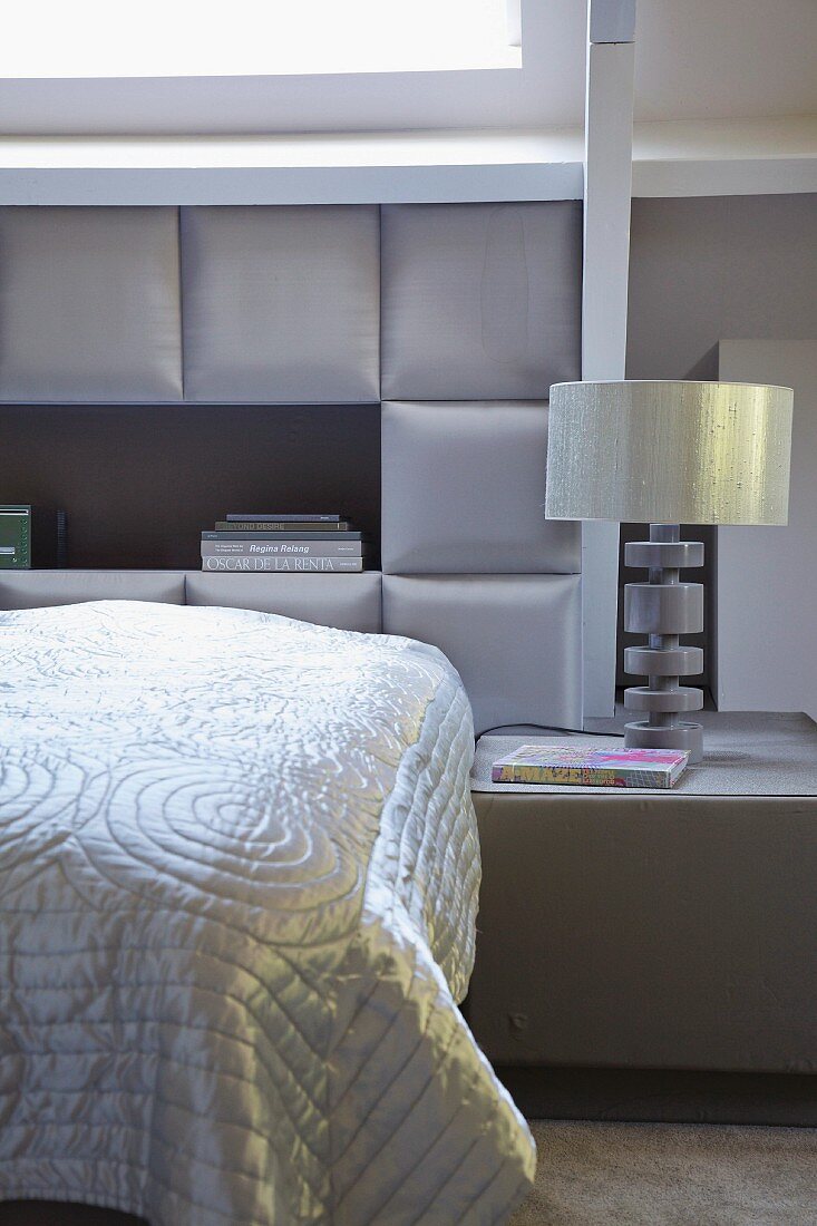 Glossy cover on bed against upholstered wall panels and table lamp on bedside table in bedroom in elegant shades of grey