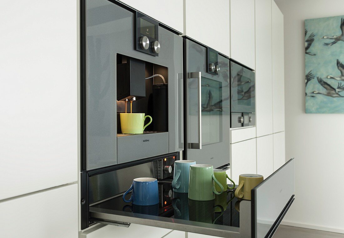 Colourful mugs on an open warming drawer below a built-in coffee machine