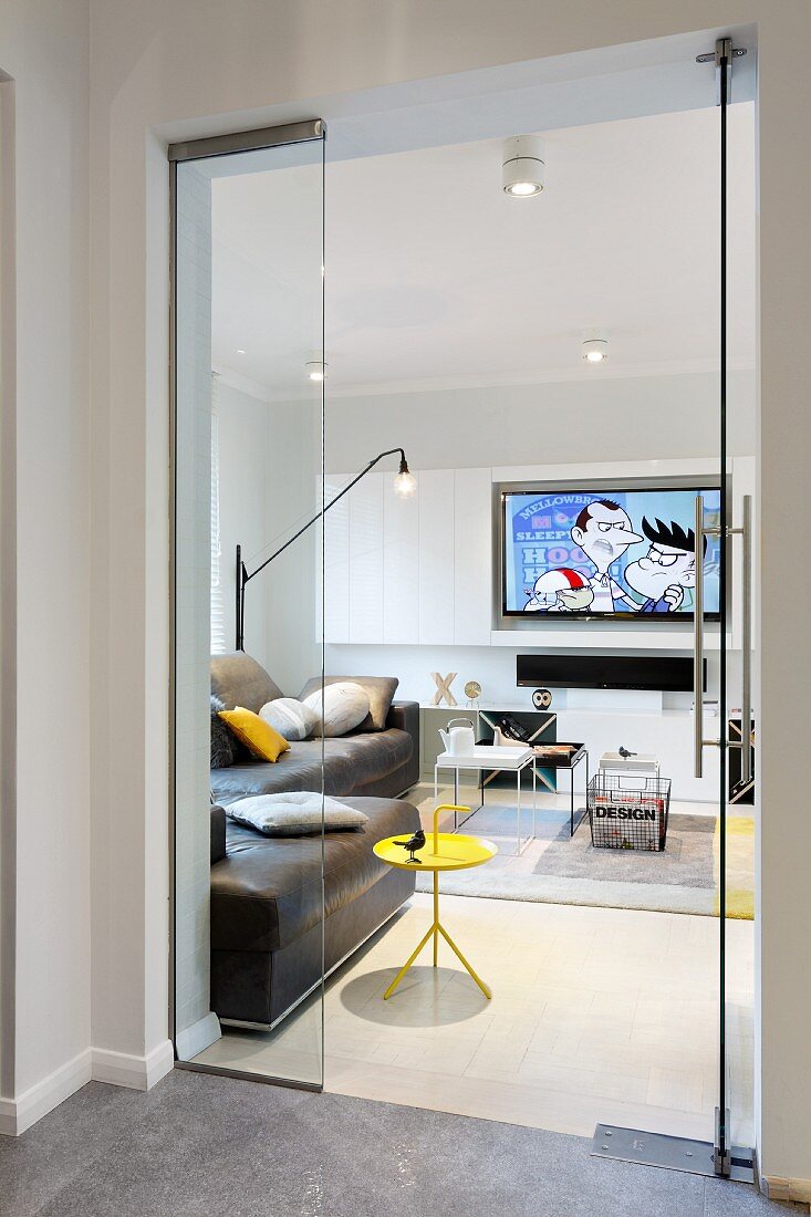 View through open glass doors of yellow side table, brown leather sofa and flatscreen TV on wall