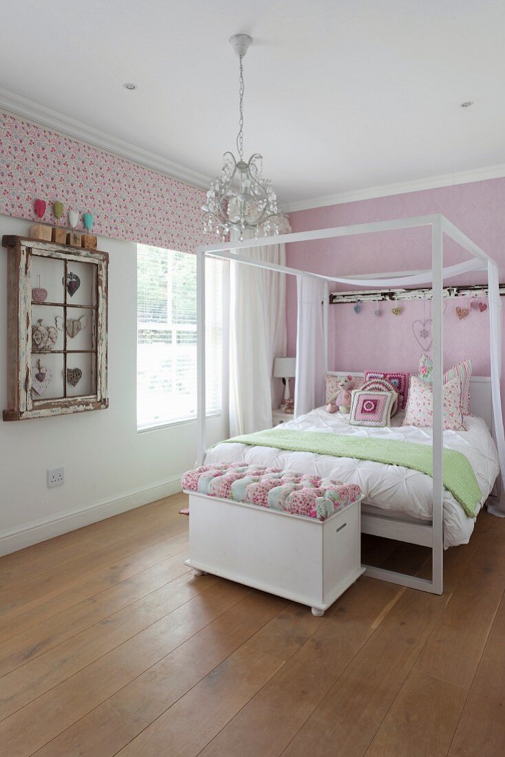 Double bed with white canopy frame and ottoman at foot on rustic wooden floor in pink-painted girl's bedroom