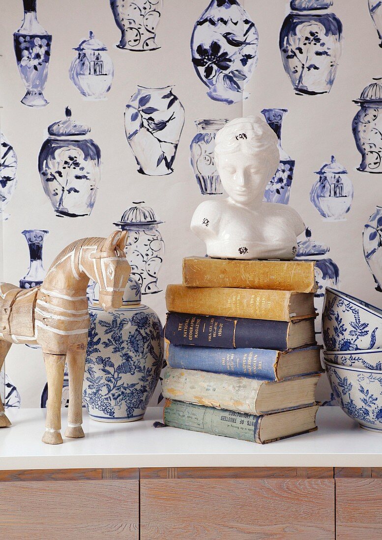 Frauenbüste auf Bücherstapel, blau-weiss bemalter Keramik Vase und Schüsseln vor blau-weisser Tapete mit Vasenmotiven