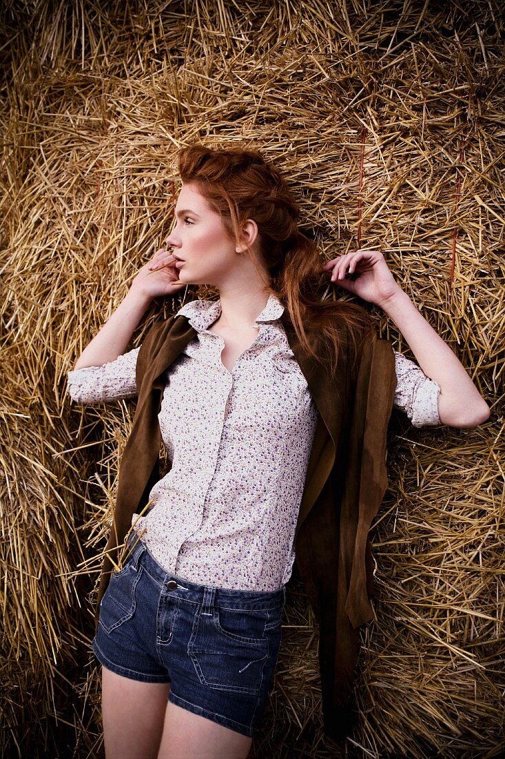 A young woman wearing denim shorts, a floral-patterned blouse and a leather gillet standing by bales of hay